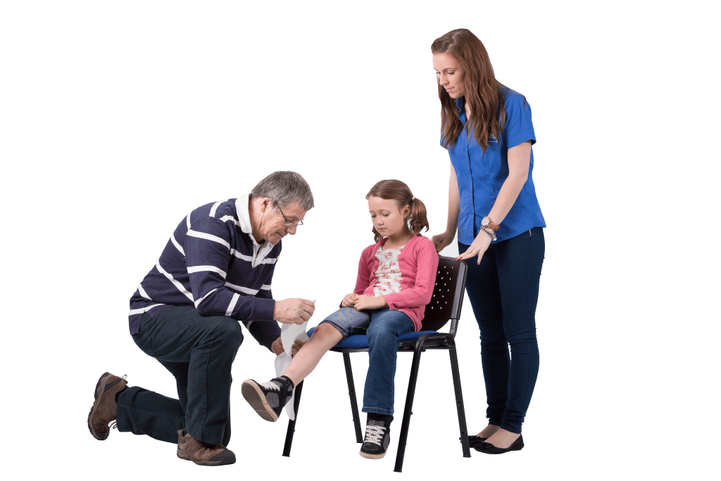 a small girl sits on a chair whilst a man bandages her right Knee. A woman is stood behind the chair overlooking the procedure.